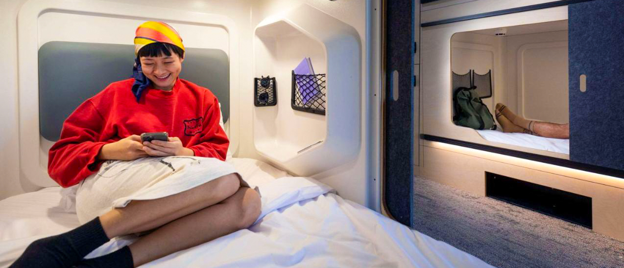 A happy traveler relaxes in a modern capsule hostel bed, smiling at her phone in Auckland.