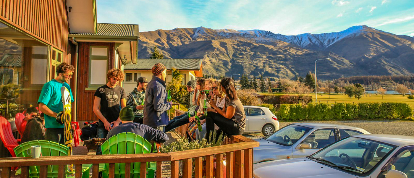 Backpackers relax on a hostel deck, enjoying drinks and conversation with snow-capped mountains in Wānaka.