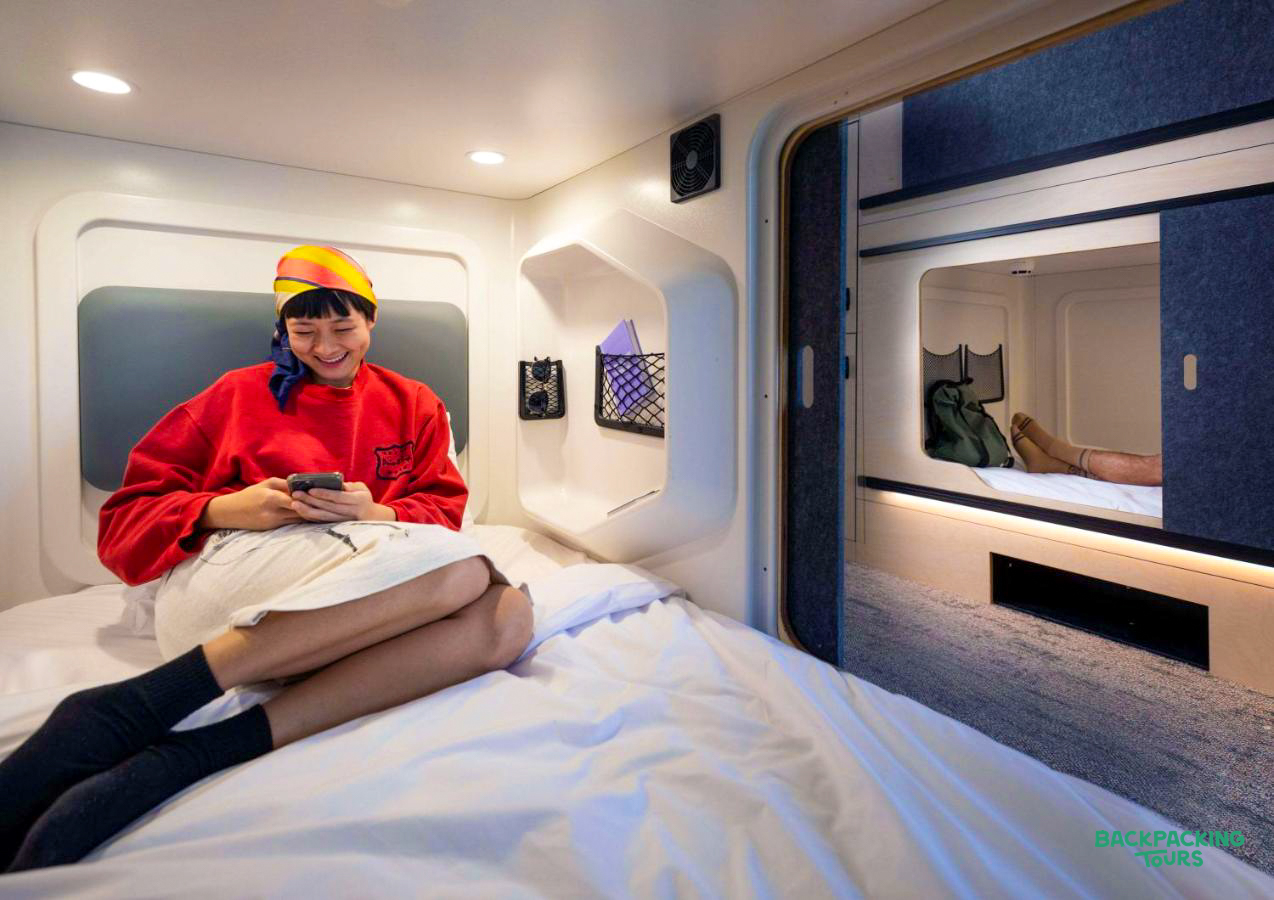 A happy backpacker relaxes on a comfortable bed in a modern Auckland hostel capsule.
