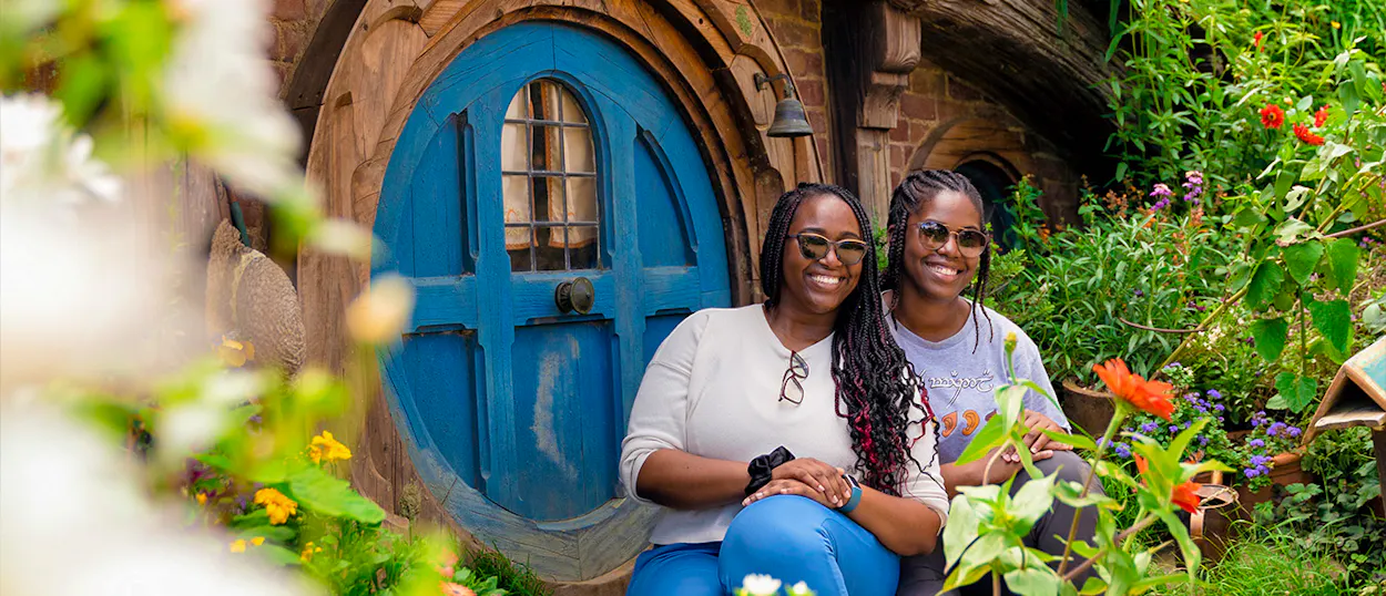 Two smiling women sit in front of a round blue door at Hobbiton, surrounded by flowers.