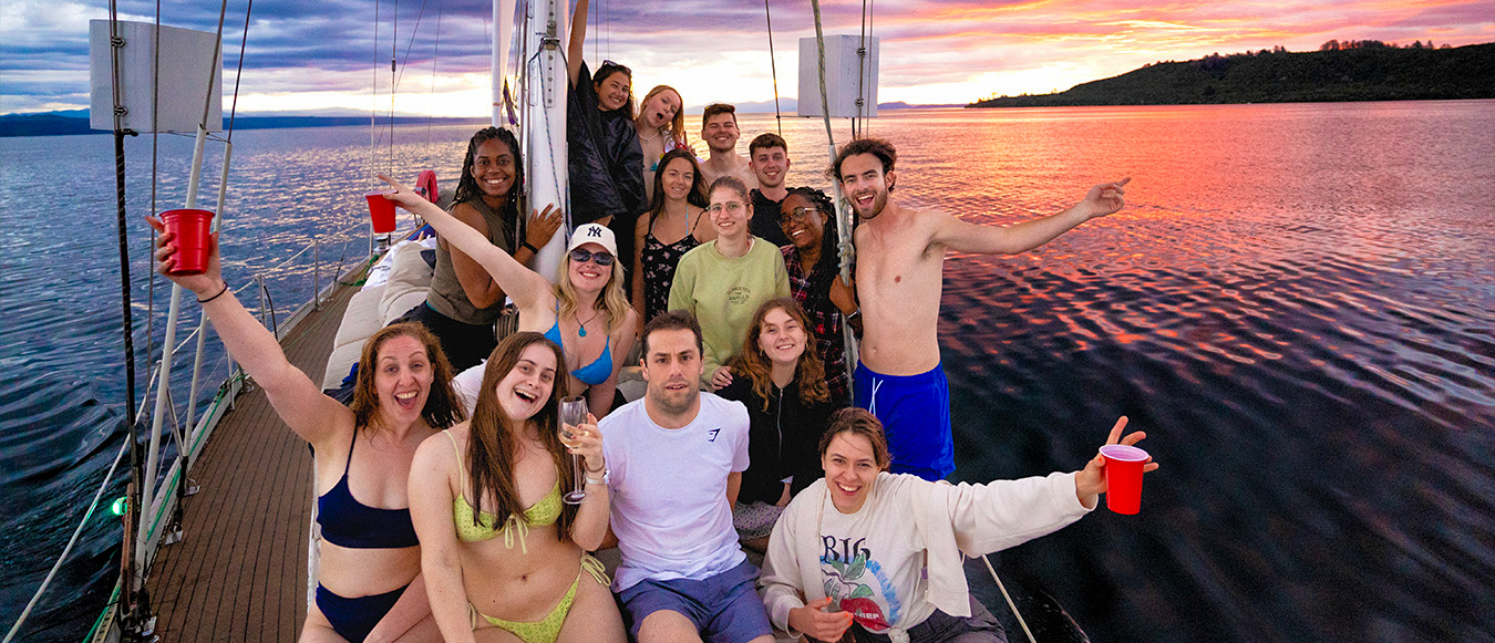 A group of happy young travelers on a boat enjoying a vibrant sunset over Lake Taupo.