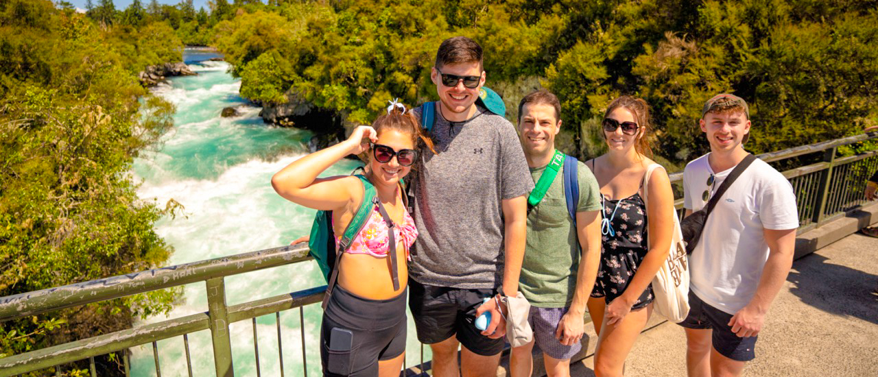 Five happy backpackers pose on a bridge overlooking the turquoise rapids of Huka Falls in New Zealand.