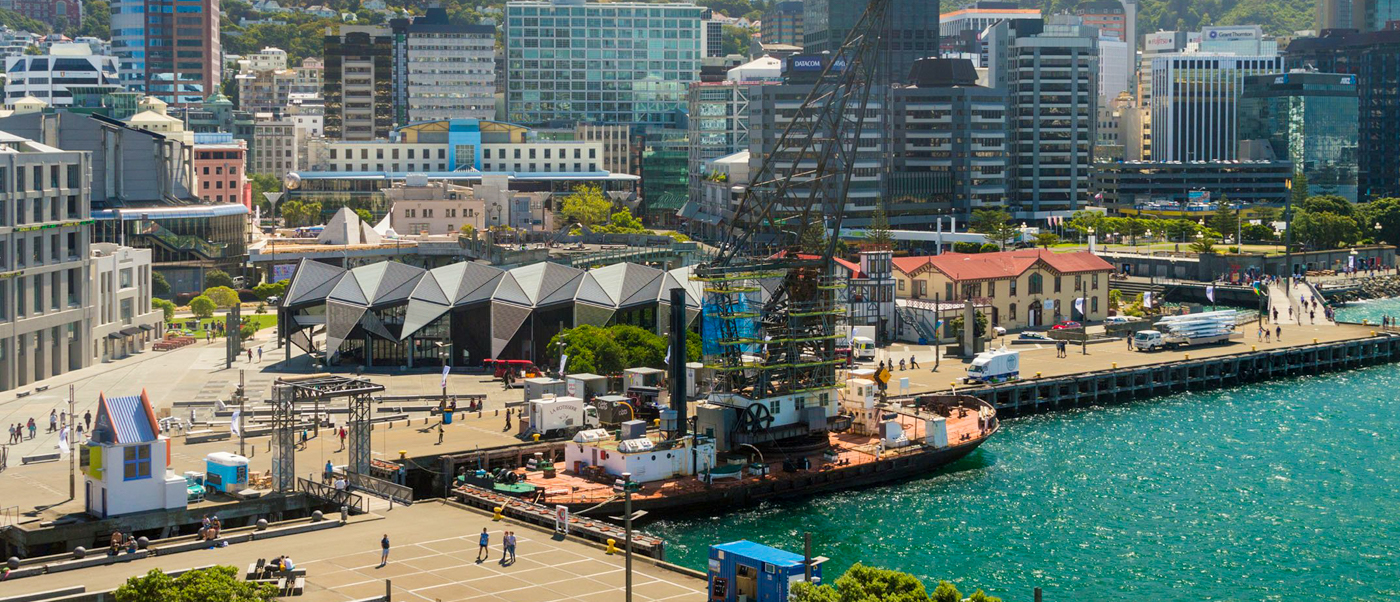 People stroll along Wellington's bustling waterfront, with a city skyline, geometric buildings, and a crane boat.
