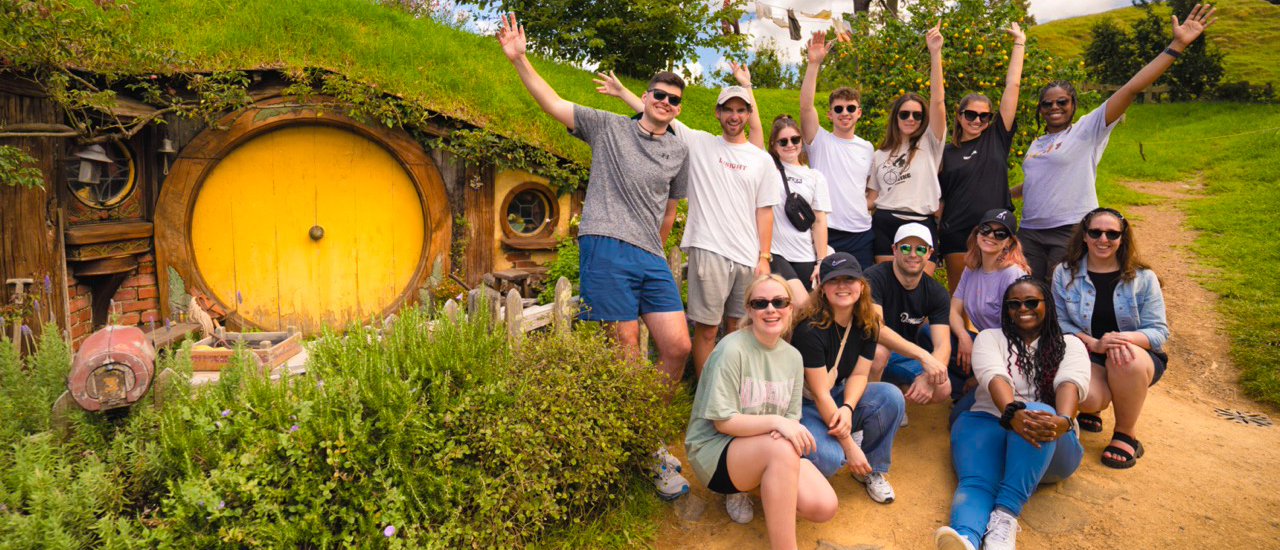 Excited backpackers pose and wave in front of a round yellow Hobbit door in Hobbiton, New Zealand.