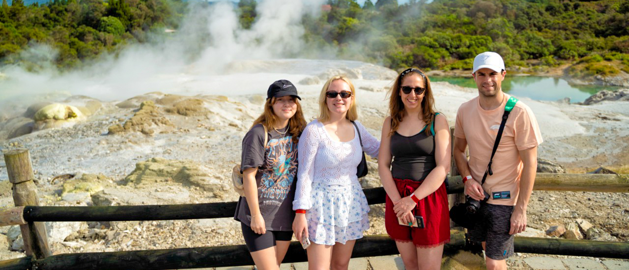 Four friends smiling in front of steaming geothermal features and vibrant pools in Rotorua, New Zealand.
