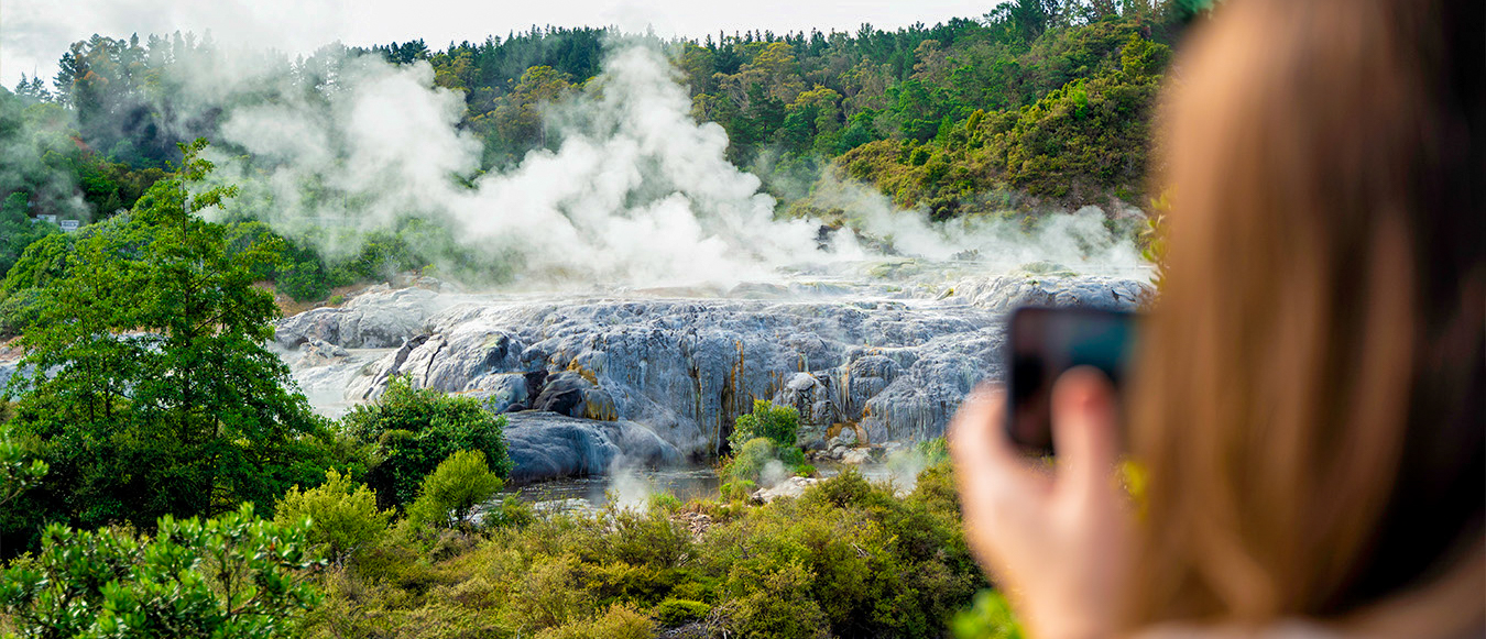 A person photographs a steamy geothermal landscape with white rock formations and green trees in Rotorua, New Zealand.