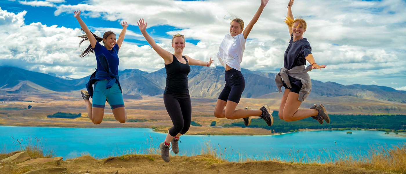 Four happy women jump high with arms raised overlooking turquoise Lake Tekapo and mountains.