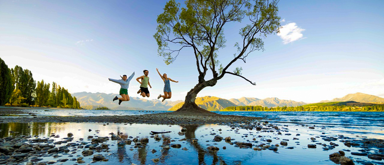 Three happy people jump in front of the iconic Wānaka tree with a lake and mountains.