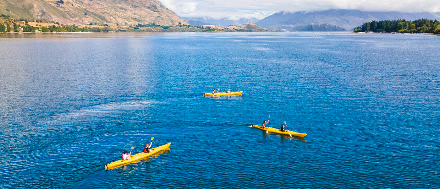 Three yellow kayaks with people paddling on the calm blue waters of Lake Wanaka, surrounded by mountains.
