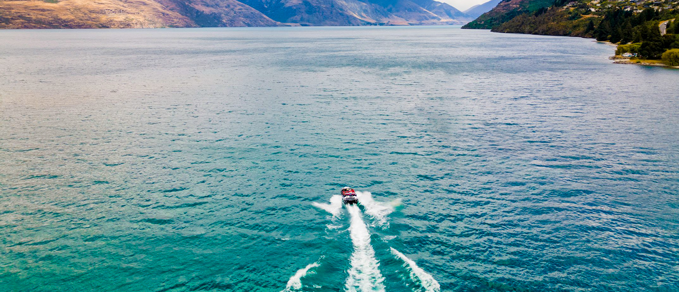 A jet boat speeds across the clear blue waters of Lake Wakatipu, with mountains in the distance.
