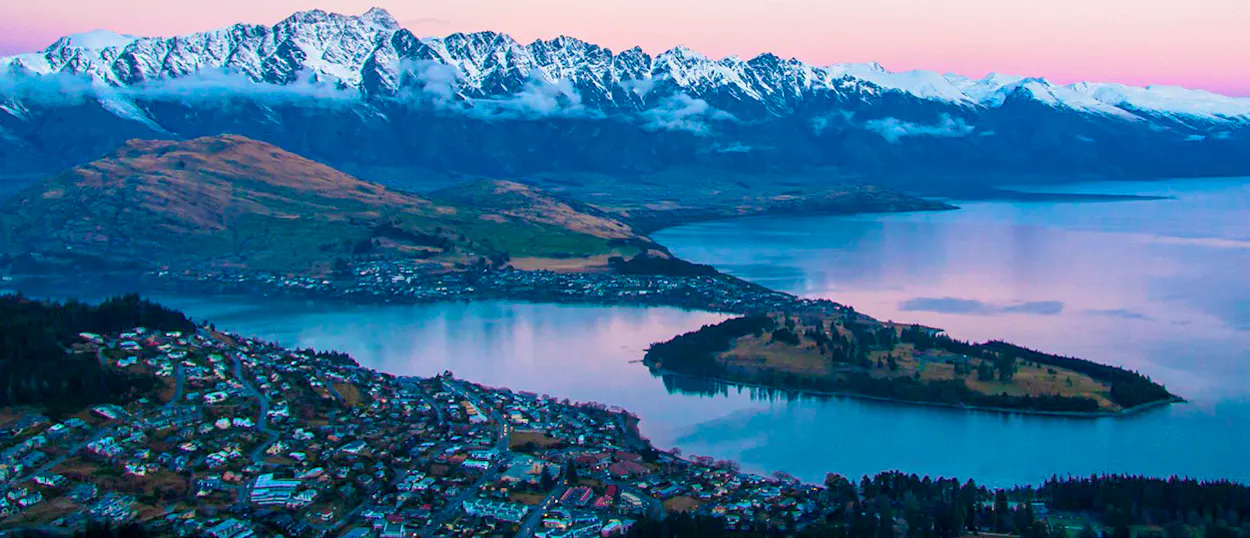 Queenstown, New Zealand, with a town, a serene lake, and majestic snow-capped mountains under a pink sky.