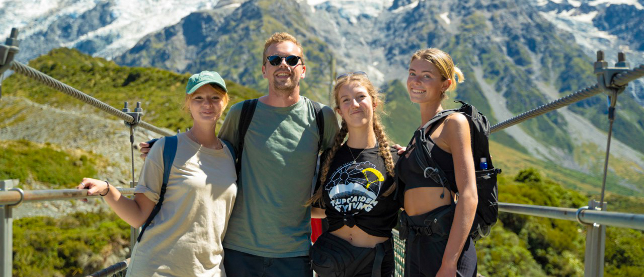 Four backpackers smiling on a swing bridge overlooking snow-capped peaks in Mount Cook National Park.