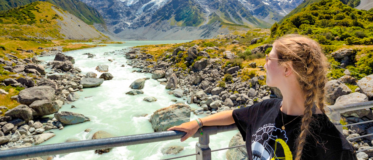 A woman on a bridge admires the turquoise glacial river and snow-capped mountains in Hooker Valley.