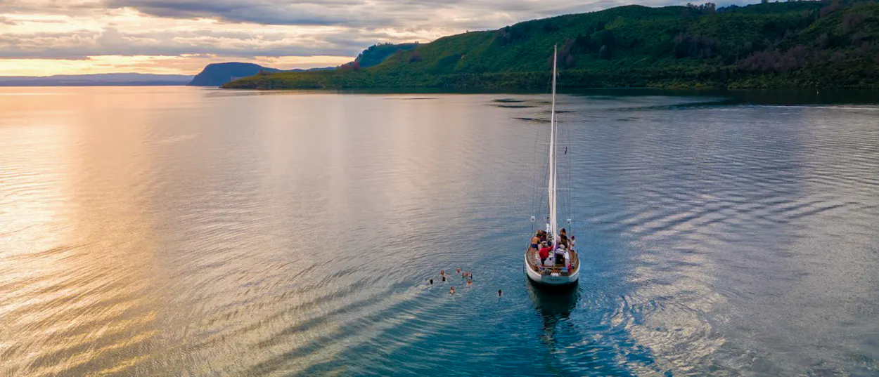 Sailboat with people cruising on calm Lake Taupo at sunset, surrounded by green hills.