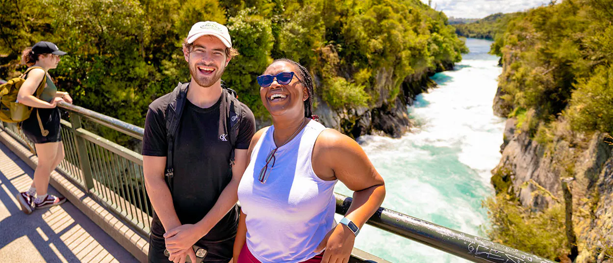 Two happy travelers laugh on a bridge overlooking the powerful turquoise Huka Falls in New Zealand.