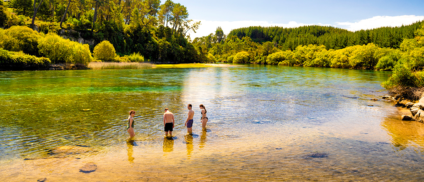 Four friends wade in the clear, green river at Taupo, surrounded by lush native bush.