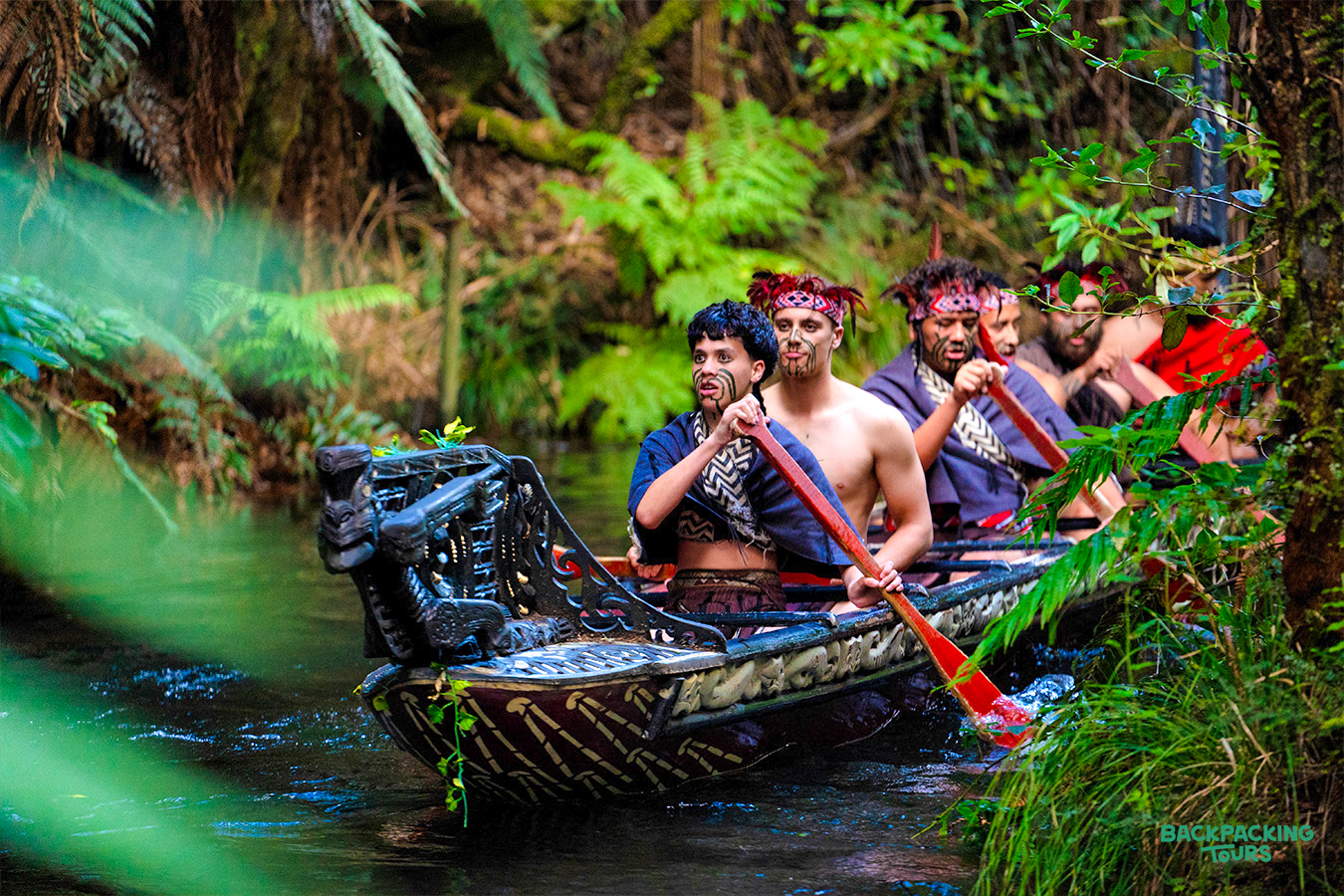 Māori men in traditional attire paddle a waka through a lush green forest in Rotorua.