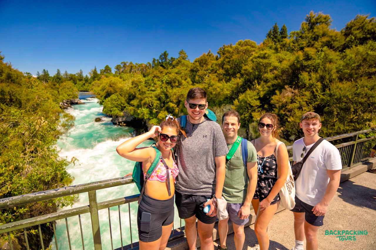 Five smiling backpackers on a bridge overlooking the powerful turquoise waters of Huka Falls, New Zealand.