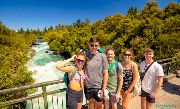 Five smiling backpackers on a bridge overlooking the powerful turquoise waters of Huka Falls, New Zealand.