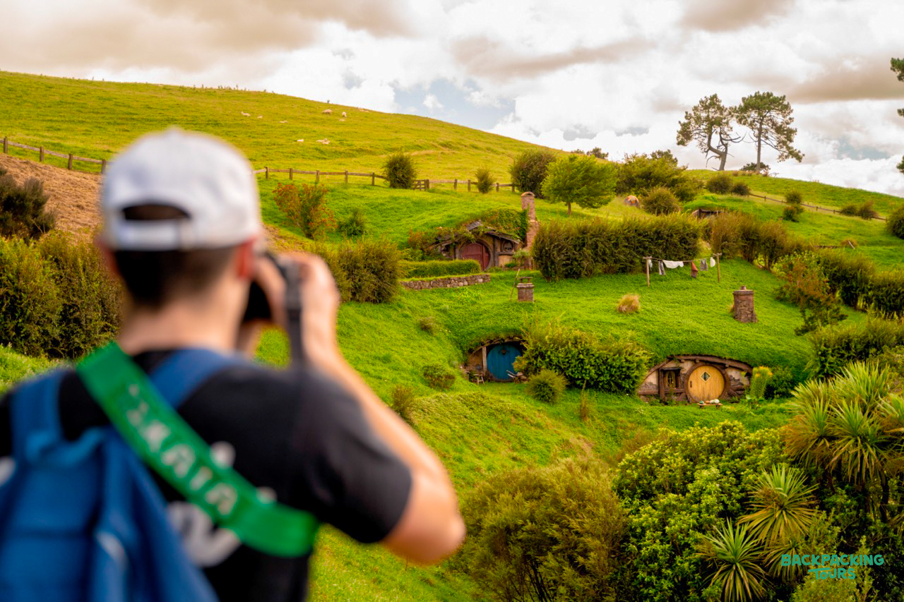 A backpacker photographs the charming hobbit holes nestled into the vibrant green hills of Hobbiton.