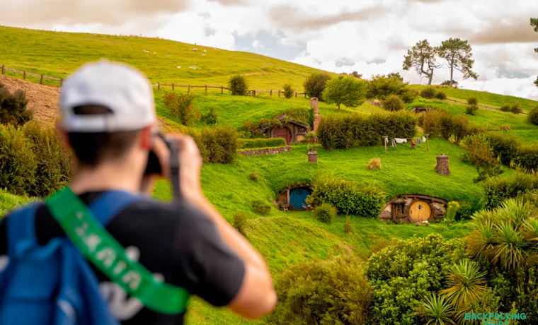A backpacker photographs the charming hobbit holes nestled into the vibrant green hills of Hobbiton.