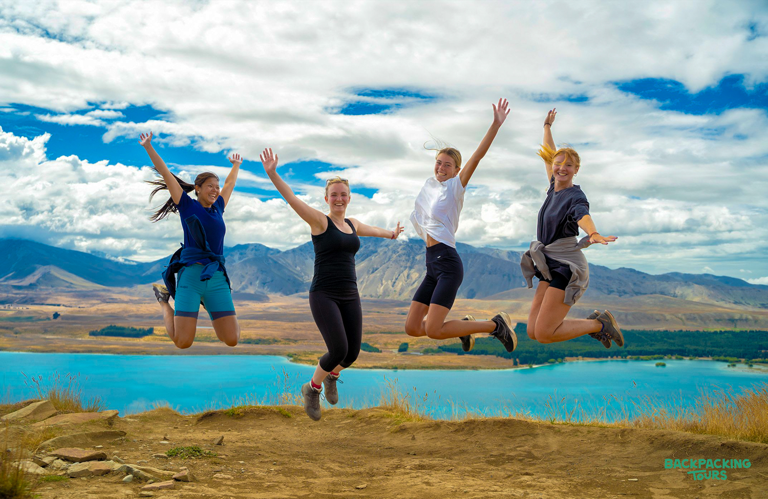 Four happy women jump with arms raised, celebrating a hike above Lake Tekapo.