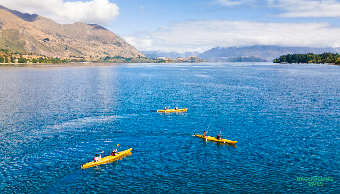 Adventurers paddle yellow kayaks on the clear blue waters of Lake Wānaka, with stunning mountains beyond.