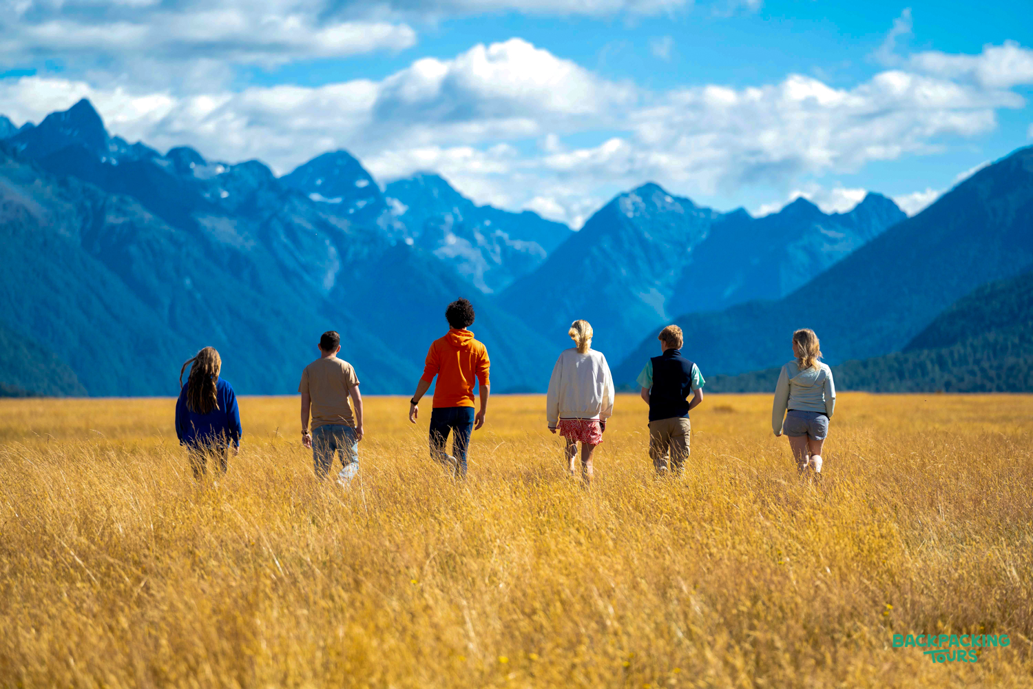 Six backpackers walk through golden grass fields with majestic blue mountains in New Zealand.