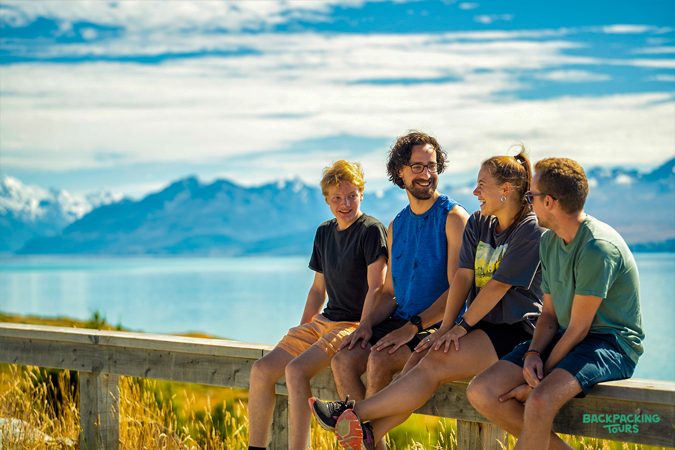 Friends share a laugh on a fence overlooking Lake Tekapo's turquoise water and snow-capped mountains.