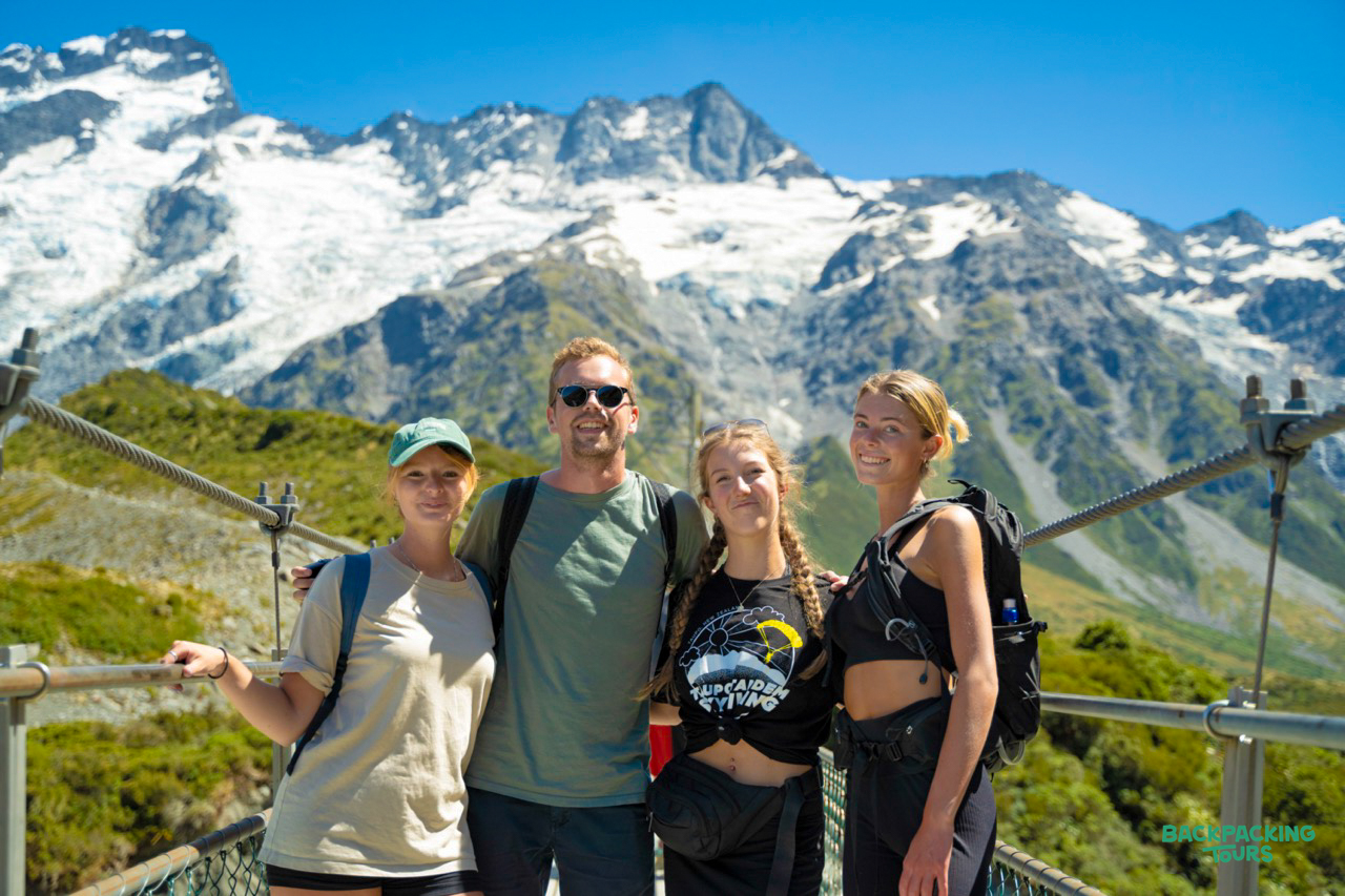 Four hikers smile on a swing bridge with snow-capped mountains in Aoraki Mount Cook National Park.