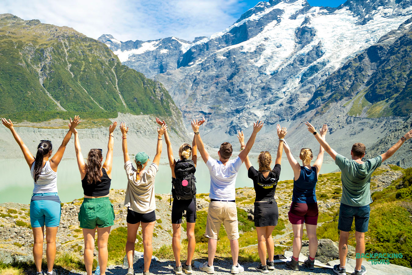 Joyful backpackers with raised arms admiring the majestic snow-capped mountains and glacial lake in Mount Cook National Park.
