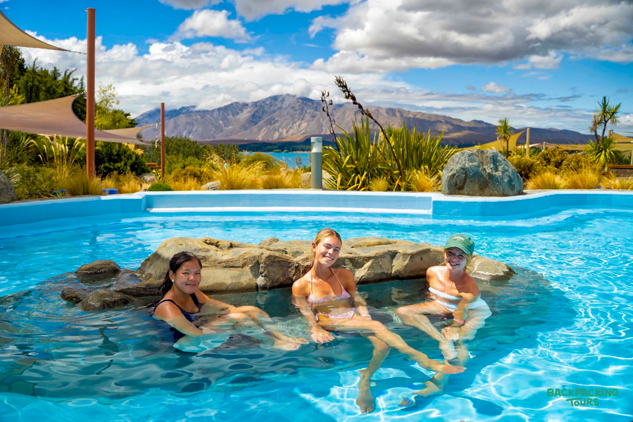Three women relax in a hot pool with stunning views of Lake Tekapo and mountains.