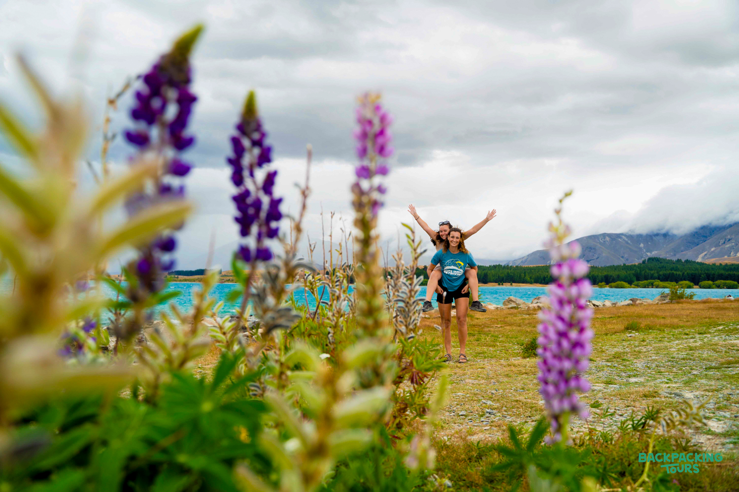 Two friends enjoy a piggyback ride by Lake Tekapo's vibrant turquoise water, framed by purple lupin flowers.