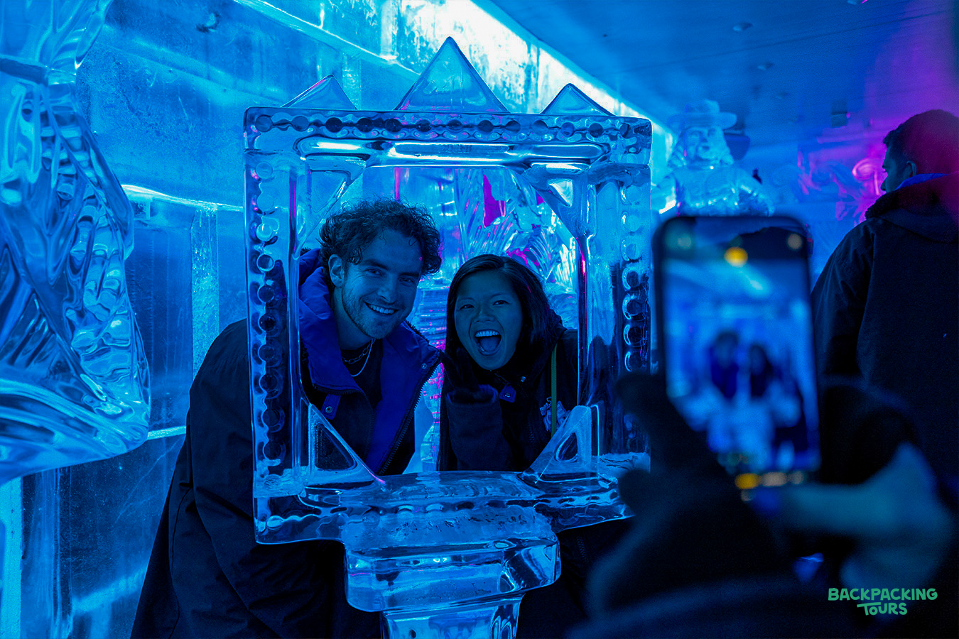 Two friends laugh and pose inside an ice frame at a vibrant blue ice bar in Queenstown.