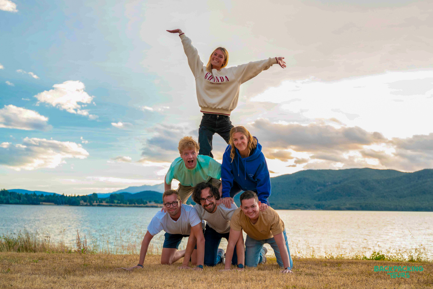 Six happy backpackers form a human pyramid by the scenic lake and mountains in Wānaka.