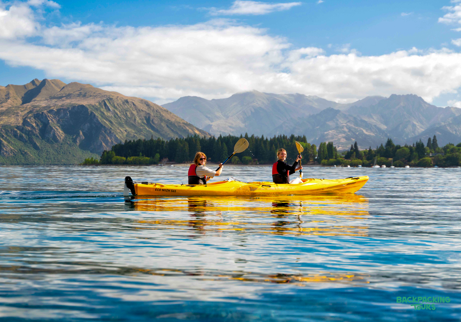 Two people paddle a yellow kayak on Lake Wānaka, New Zealand, with scenic mountains behind them.