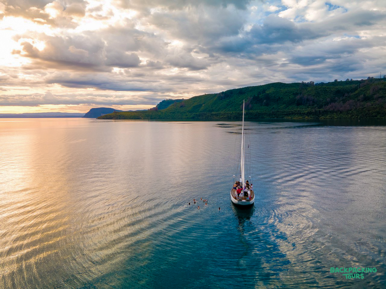 A sailboat with people on board cruises calm Lake Taupo at sunset, surrounded by green hills.