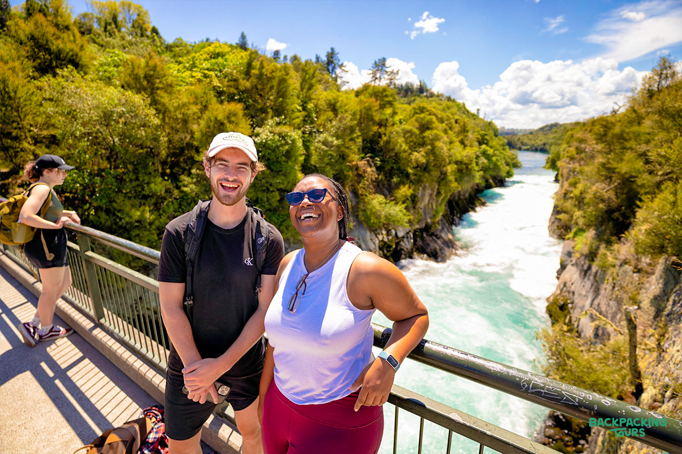 Two happy backpackers smile on a bridge overlooking the powerful Huka Falls in Taupo, New Zealand.