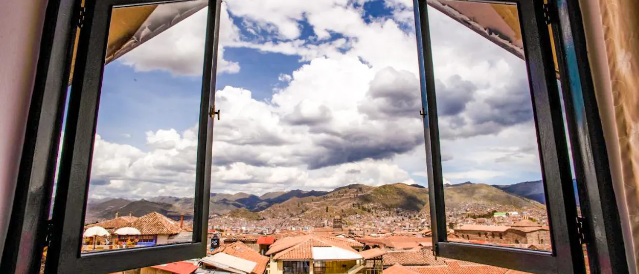 An open window frames a panoramic view of Cusco city with terracotta roofs and mountains under a cloudy sky.