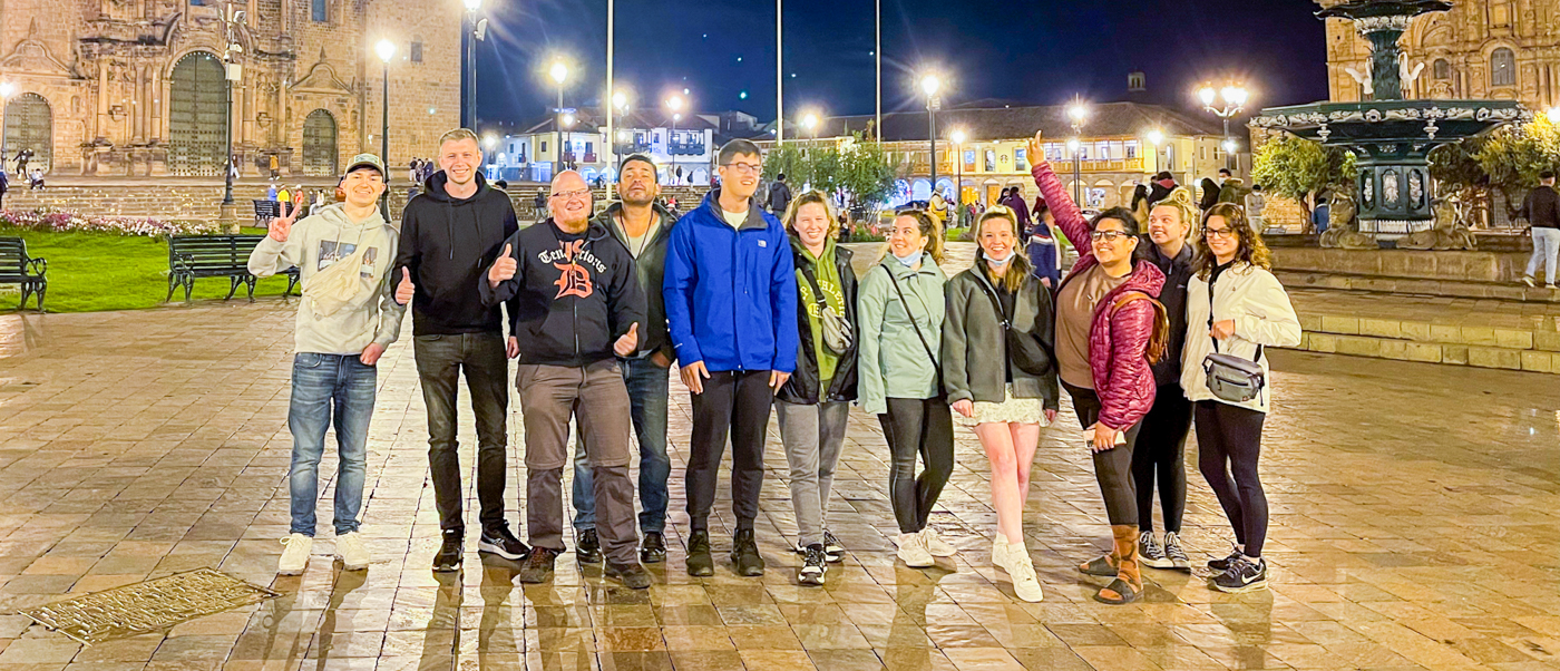 A group of happy backpackers poses together in a brightly lit historic plaza in Cusco.