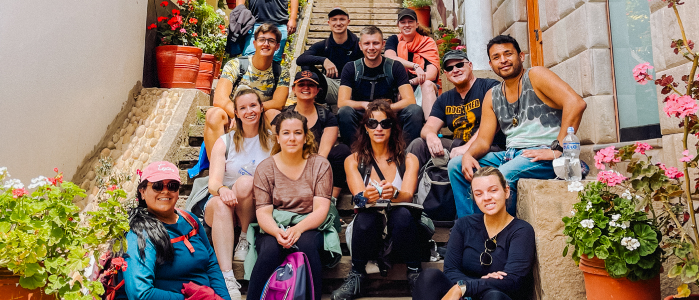 A happy group of backpackers poses on stone steps adorned with colorful flowers in Cusco.