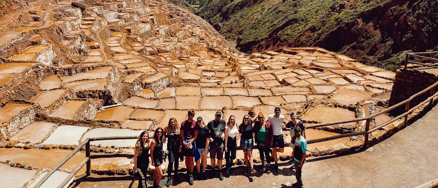 A group of happy travelers poses in front of the vast terraced Maras Salt Mines in Peru.