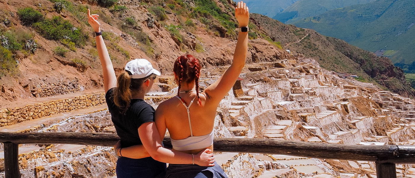 Two women with raised arms embrace, enjoying the vast Maras Salt Mines in Peru.