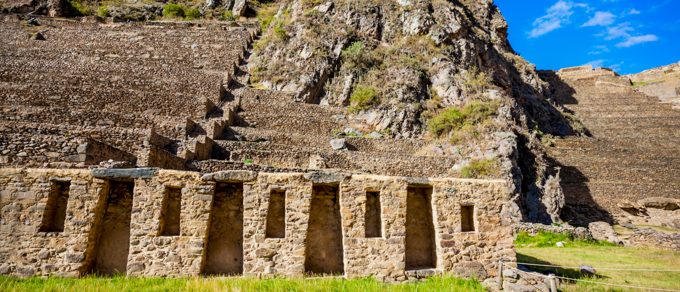 Historic Inca stone structures and agricultural terraces rise from green grass at Ollantaytambo.
