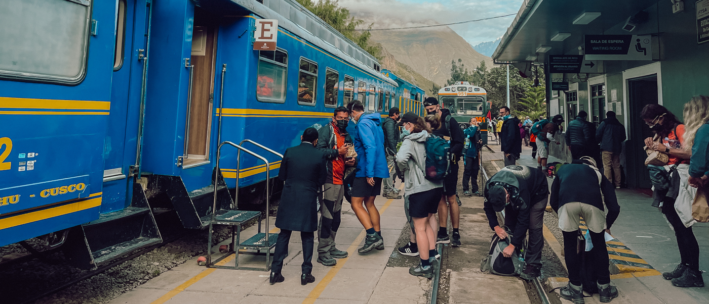 Backpackers board a blue train at a station platform, heading to Cusco, with mountains behind.
