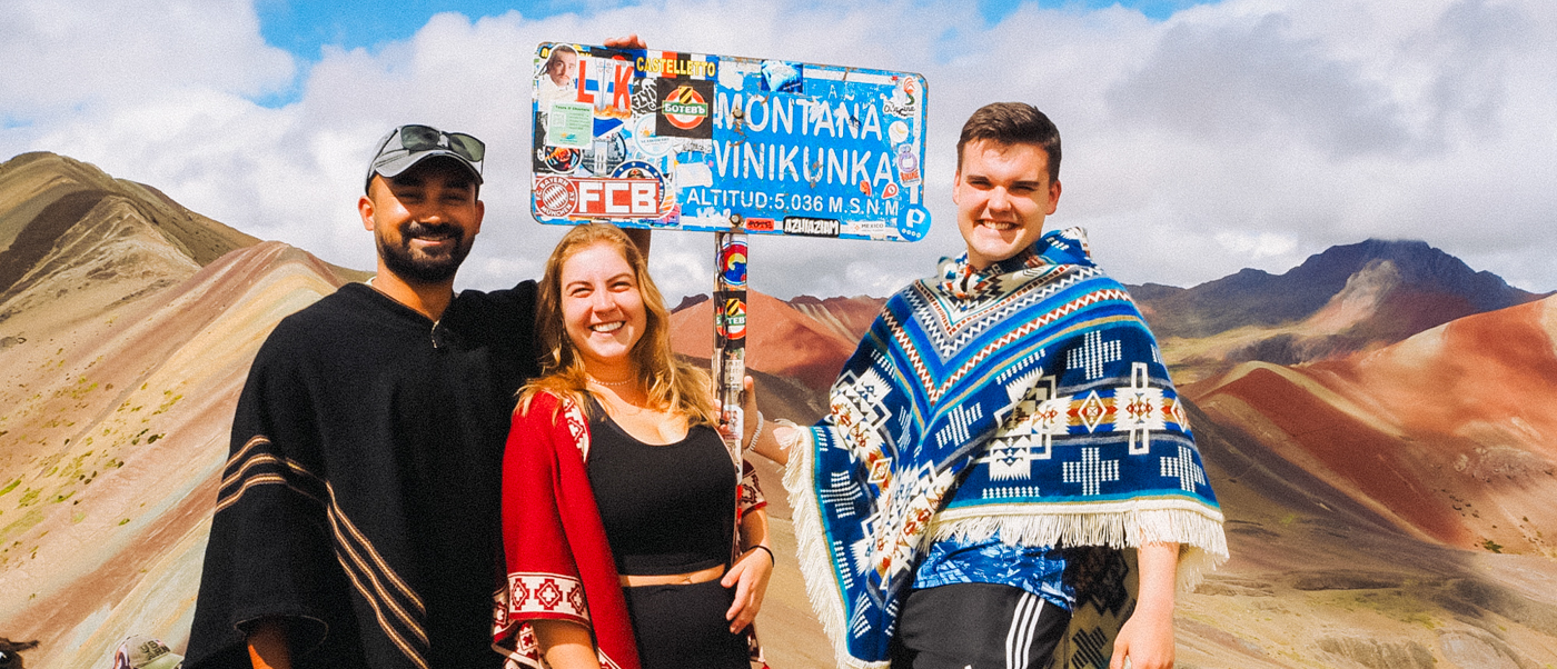 Three smiling travelers pose with the Montaña Vinicunca sign and colorful Rainbow Mountain.