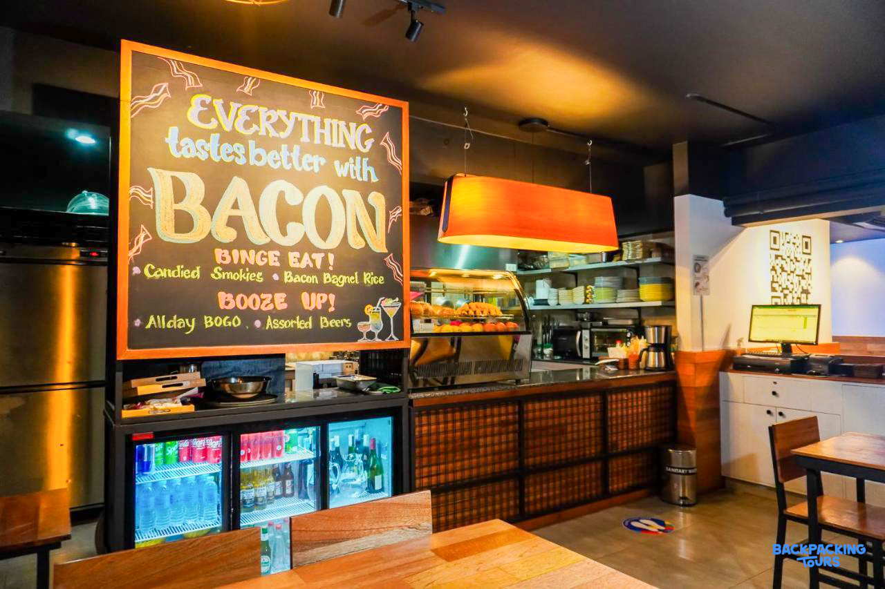 A cozy hostel cafe with a large blackboard sign promoting bacon, drinks, and a casual dining area.