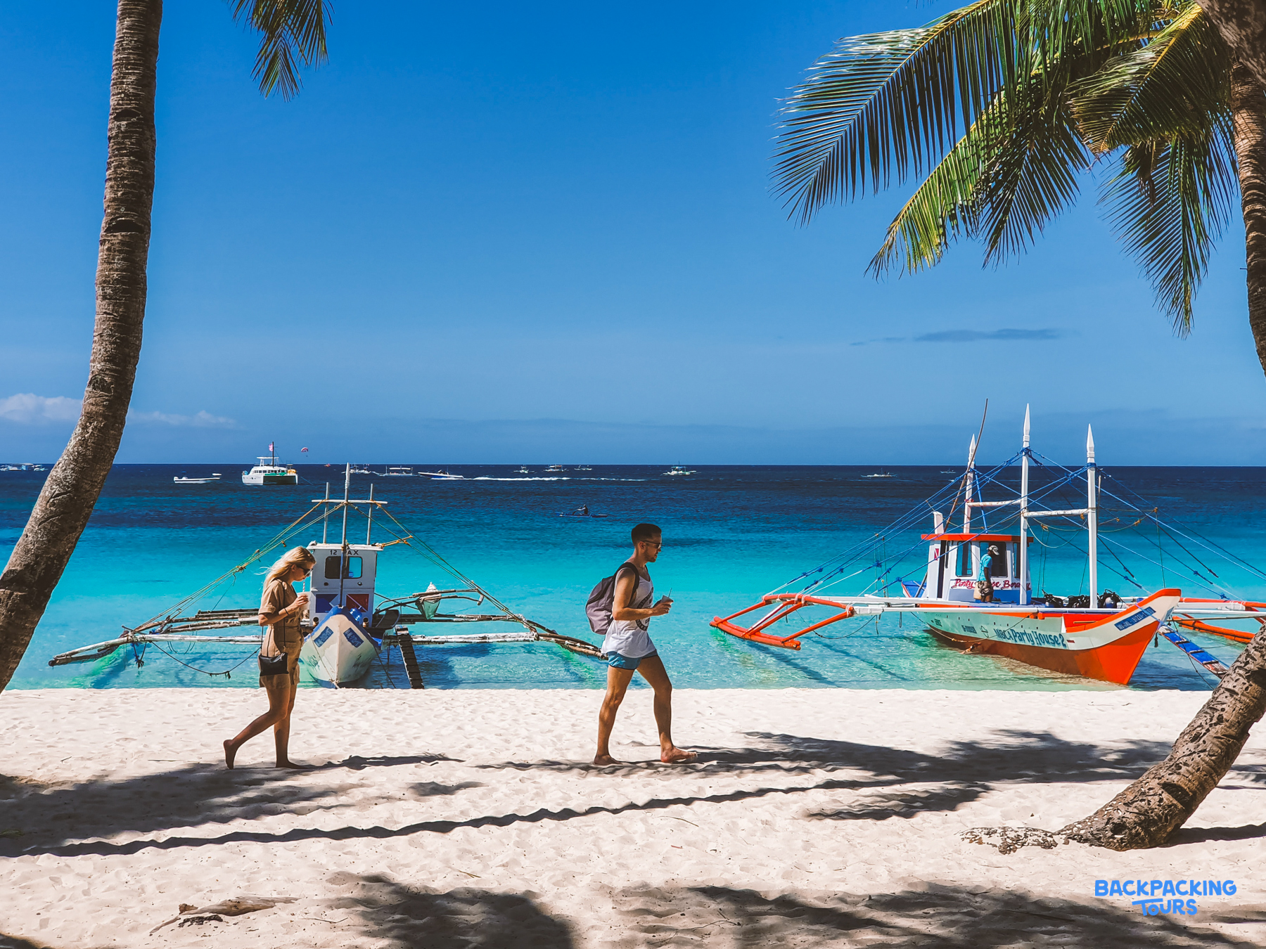 Two people walk on a beautiful white sand beach with clear turquoise water and traditional outrigger boats.