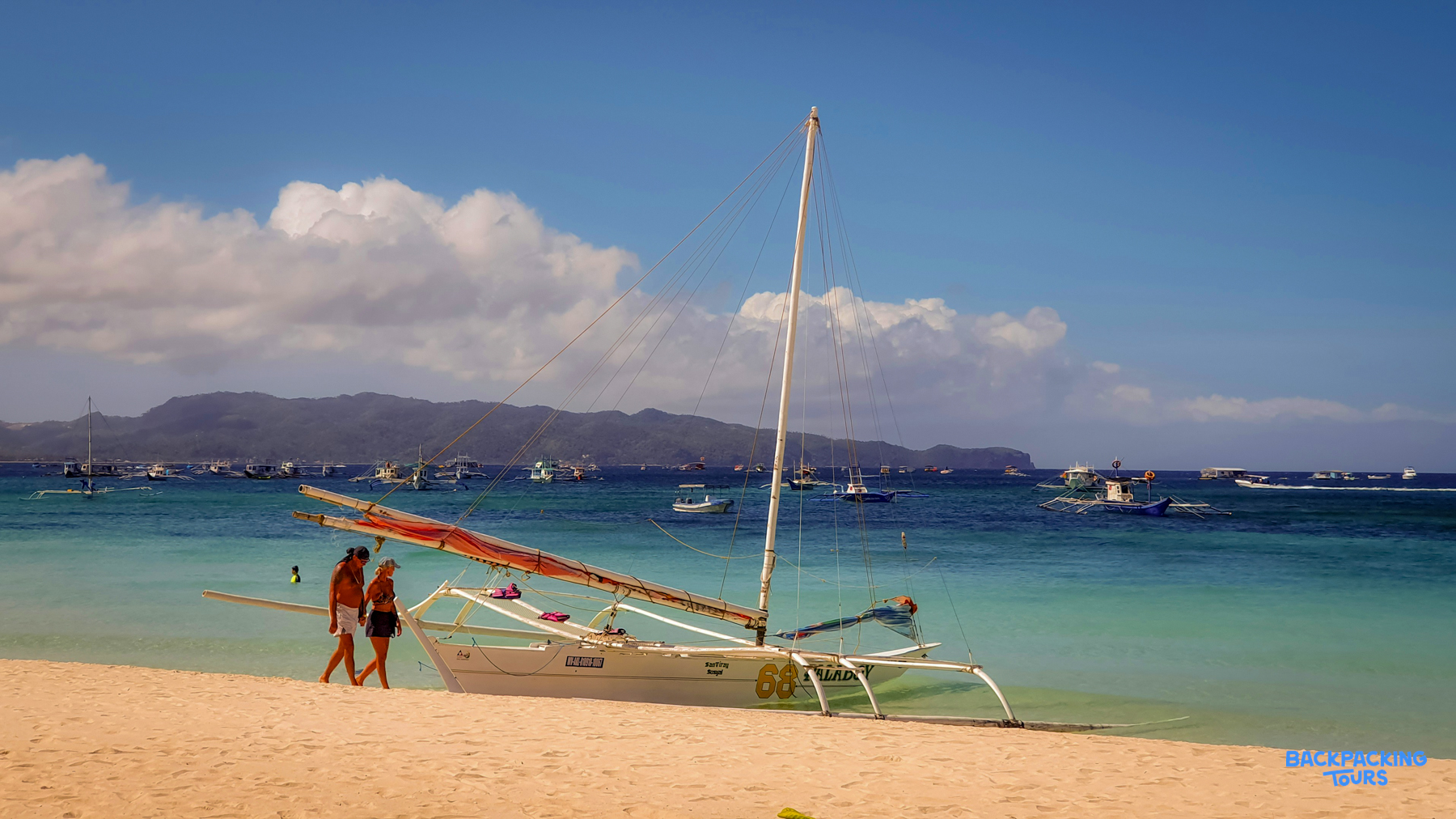 Two people stroll past a traditional outrigger boat on a beautiful white sand beach in Boracay.