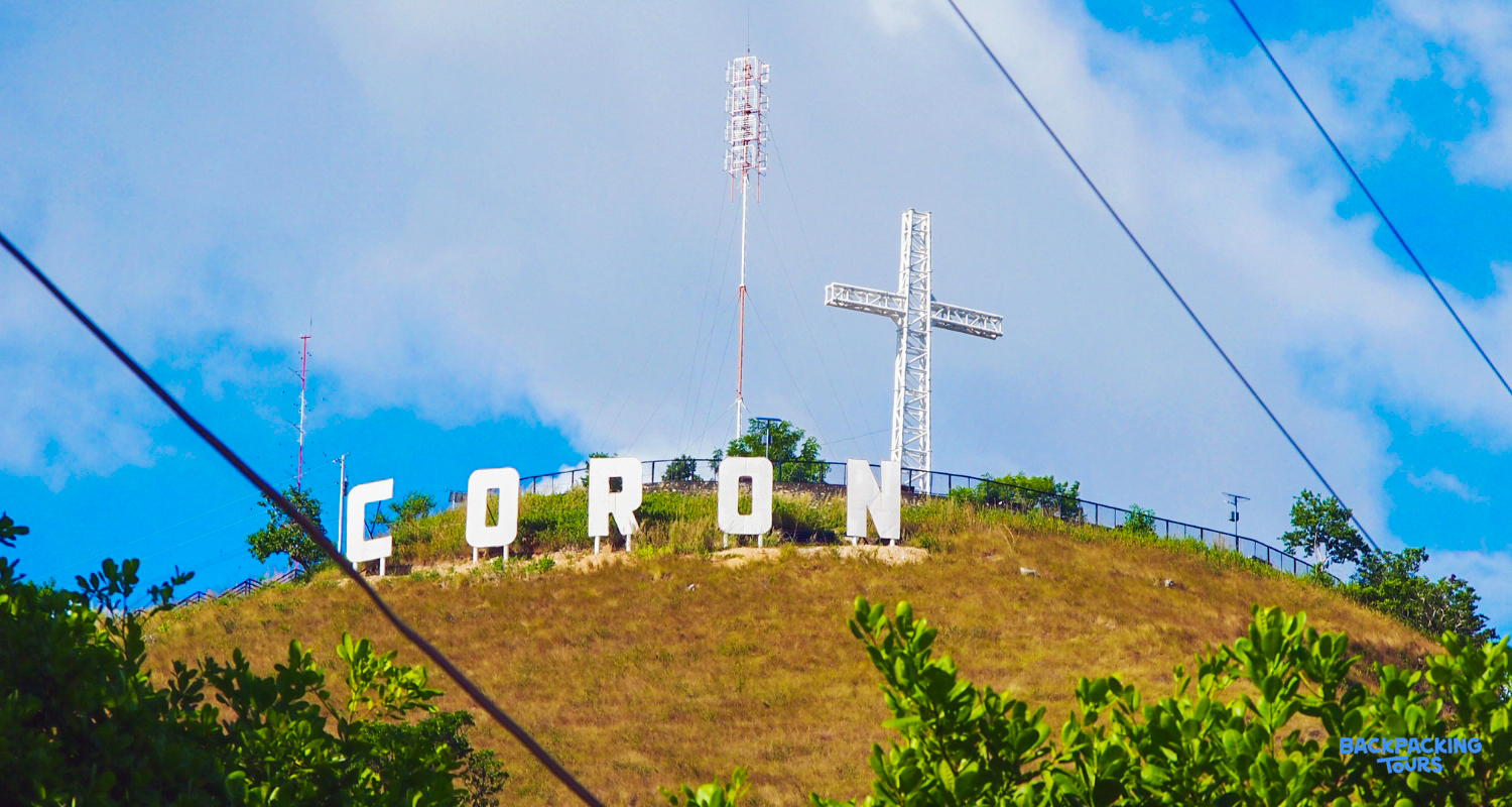 The iconic Coron sign and large white cross stand proudly atop Mount Tapyas under a clear blue sky.